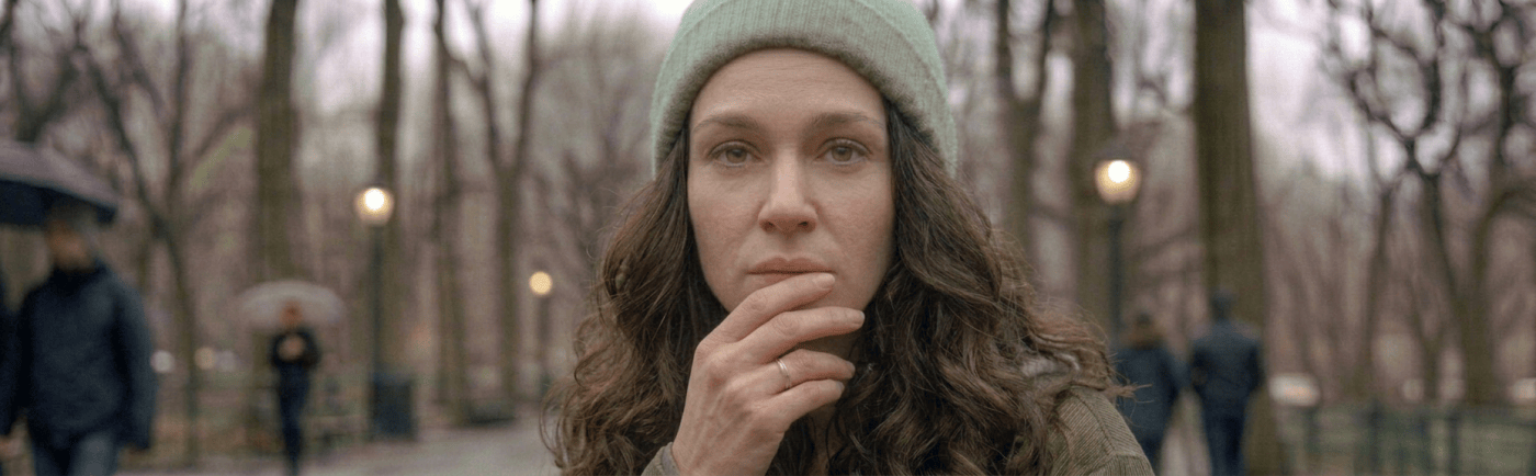 Person with gray beanie and green jacket sitting on a wet park bench, holding a cigarette, laughing with tears in their eyes.