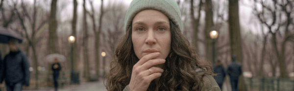 Person with gray beanie and green jacket sitting on a wet park bench, holding a cigarette, laughing with tears in their eyes.